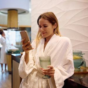 Happy woman in bathrobe using smart phone while drinking water at wellness center. Happy woman texting on cell phone while spending her weekend at health spa.